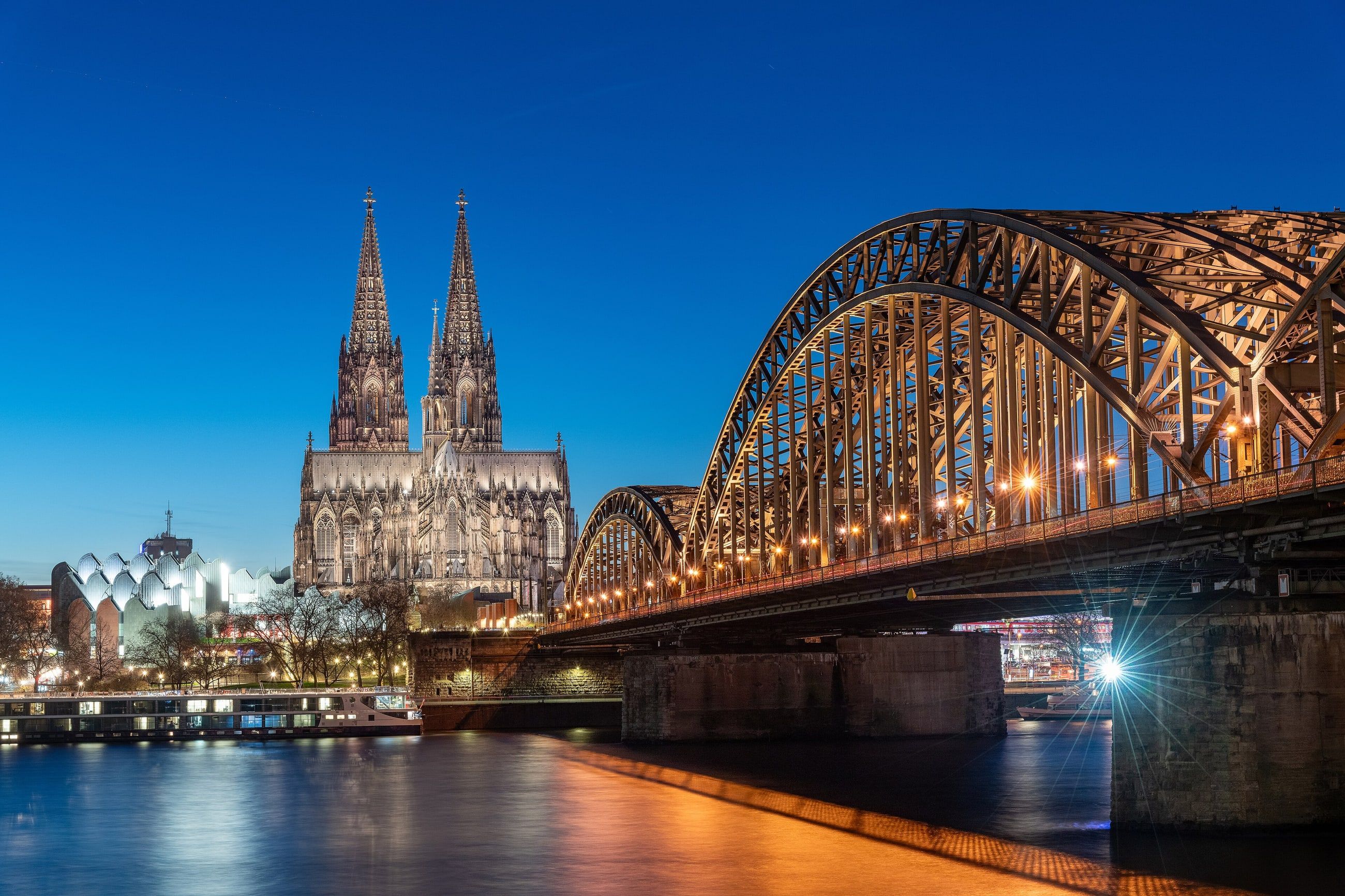 Der Kölner Dom und die beleuchtete Hohenzollernbrücke spiegeln sich zur Blauen Stunde im Rhein.
