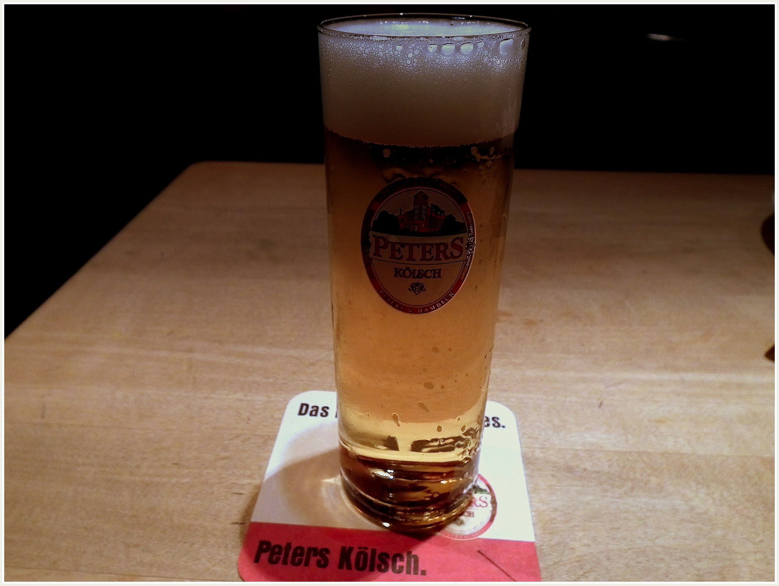 Ein Glas Kölsch in der Stange mit Schaumkrone steht auf einem Bierdeckel auf einem Holztisch.
